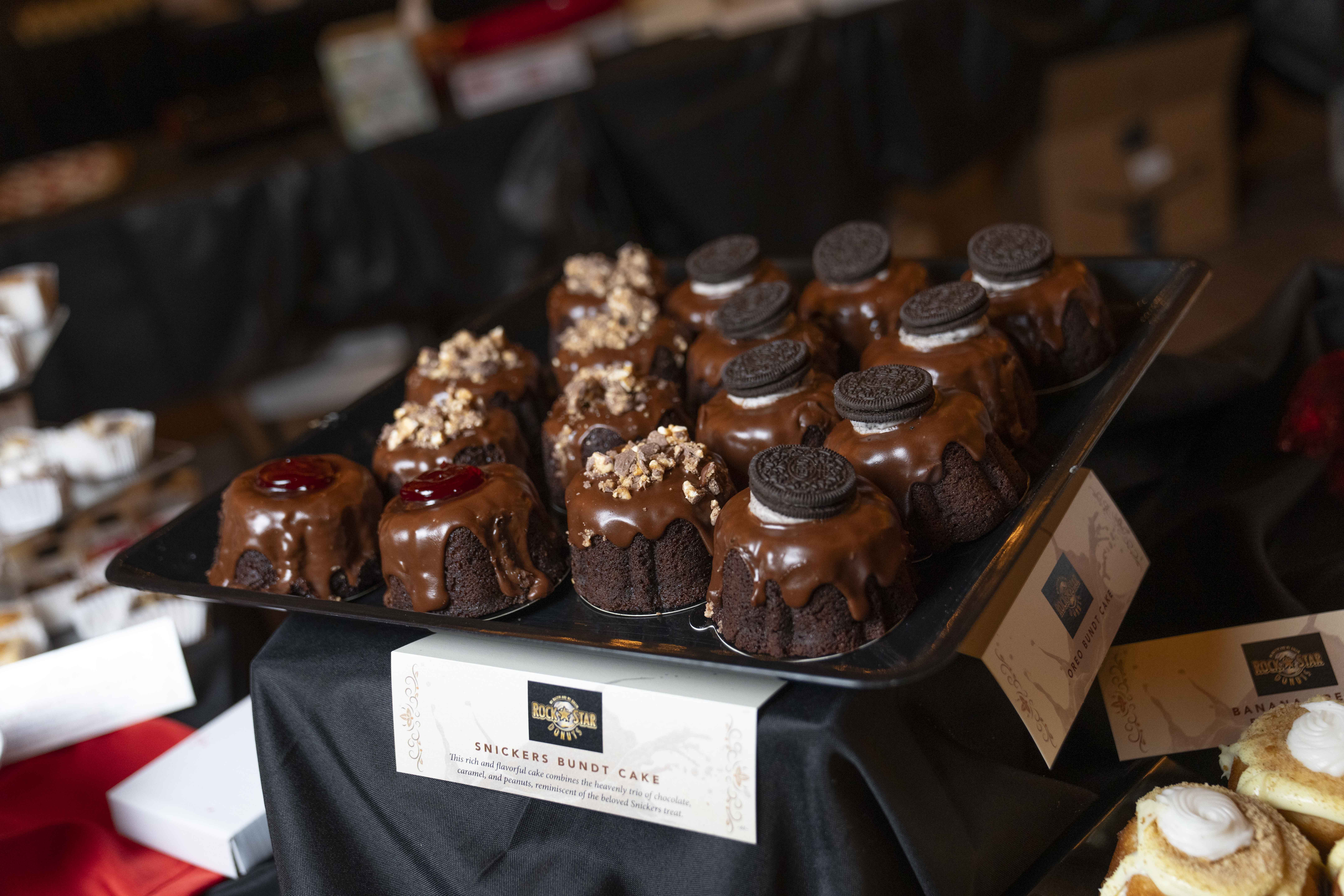 Close-up of Snickers Bundt Cake from Rock Star Donuts. Card beneath dessert reads, Snickers Bundt Cake. This rich and flavorful cake combines the heavenly trio of chocolate, caramel, and peanuts, reminiscent of the beloved Snickers treat.