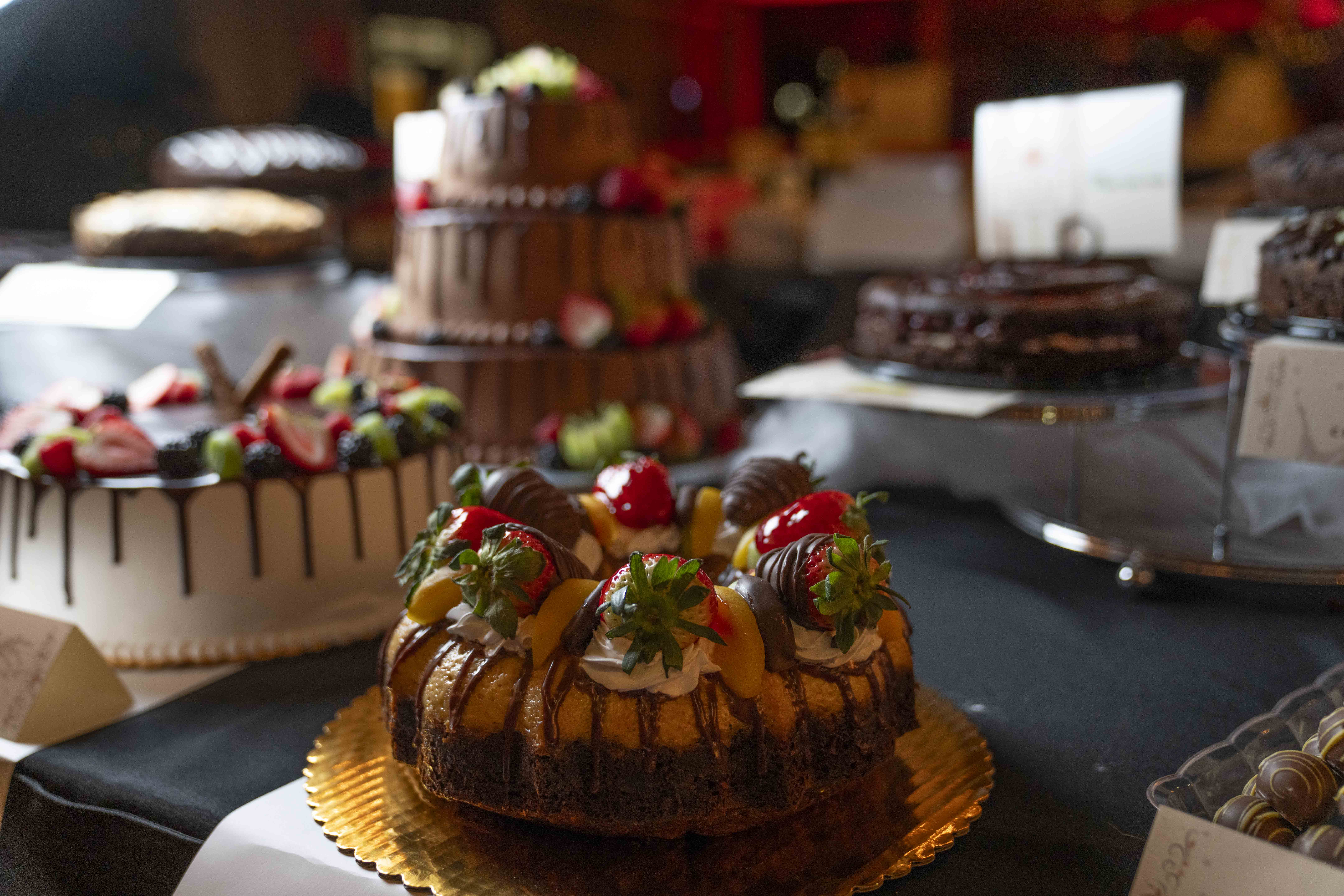 Close-up of a bundt cake with chocolate-covered strawberries on top. In the background are more cakes of varying flavors.